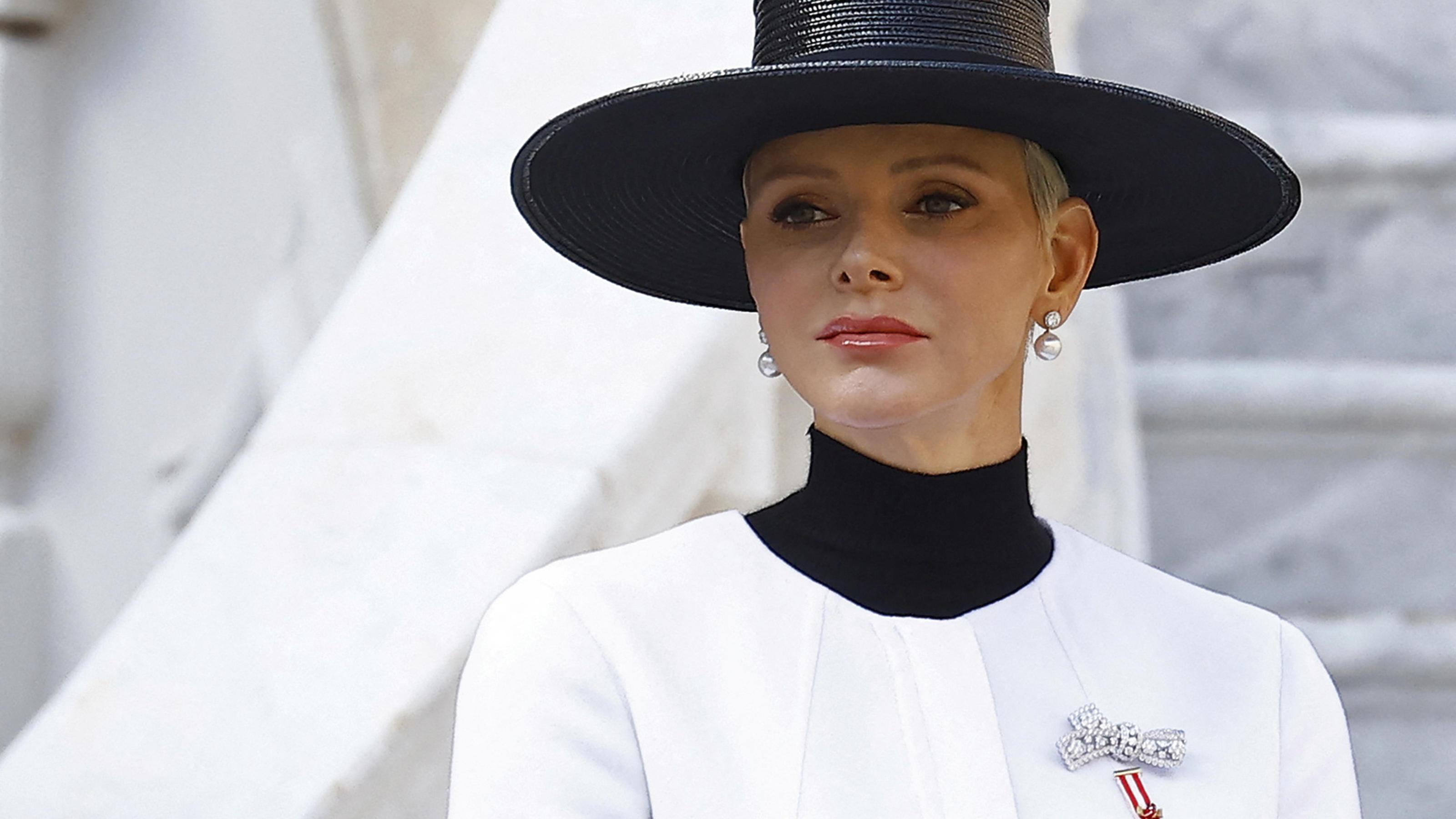 Charlène von Monaco trägt einen schwarzen Hut und ein weißes Kostüm. Sie steht vor einer Mamor-Treppe und schaut ernst zur Seite.